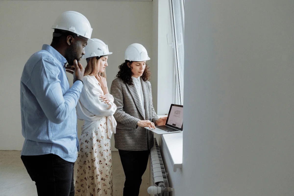 A group of three individuals in hard hats collaborate while reviewing a laptop in a bright, modern workspace.