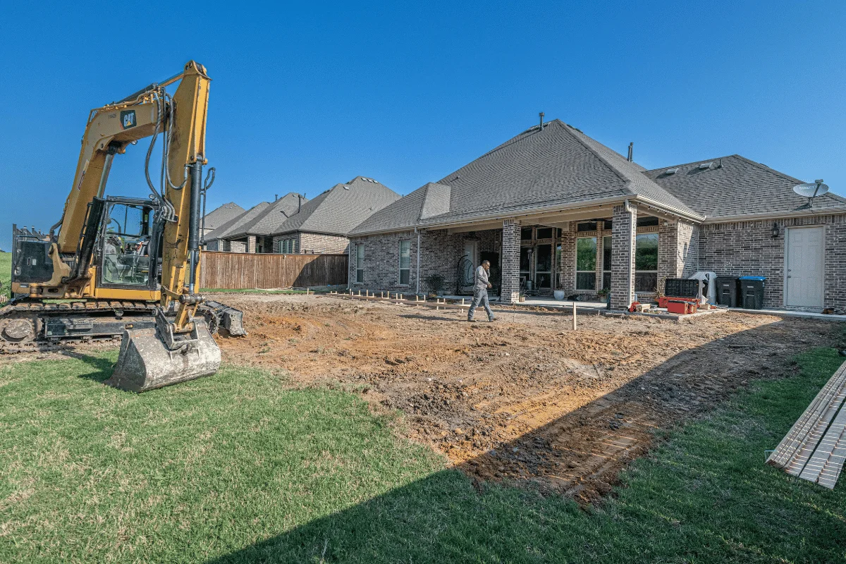 Construction scene in a backyard with an excavator, gravel ground, and a worker near a house under a clear blue sky.