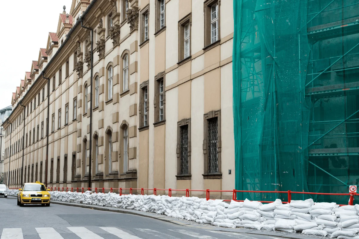 Yellow taxi and white car drive past a historic building, partially covered with green mesh, and sandbags along the street for protection.
