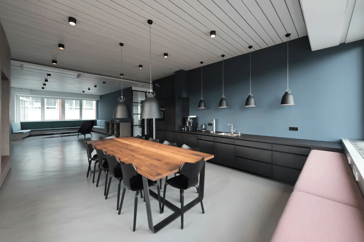 A modern kitchen and dining area with sleek black cabinets, pendant lights, and a long wooden dining table surrounded by chairs.