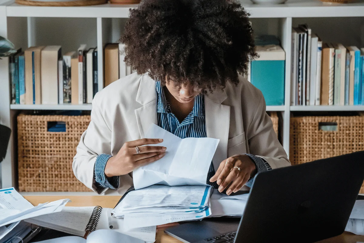 A person with curly hair in a blazer reviews documents at a cluttered desk with a laptop and bookshelves, conveying focus and concentration.
