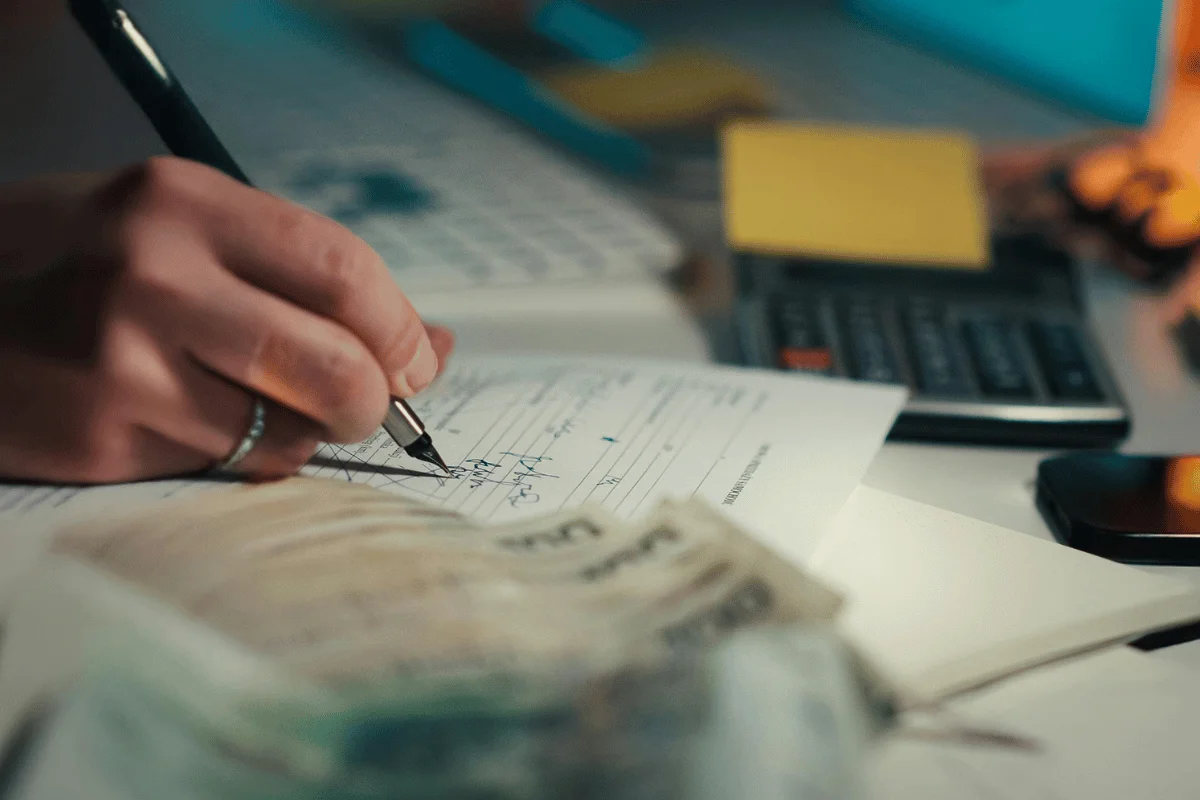 A person writes notes on paper at a cluttered desk with a calculator and scattered currency. The scene conveys a focused, financial task.