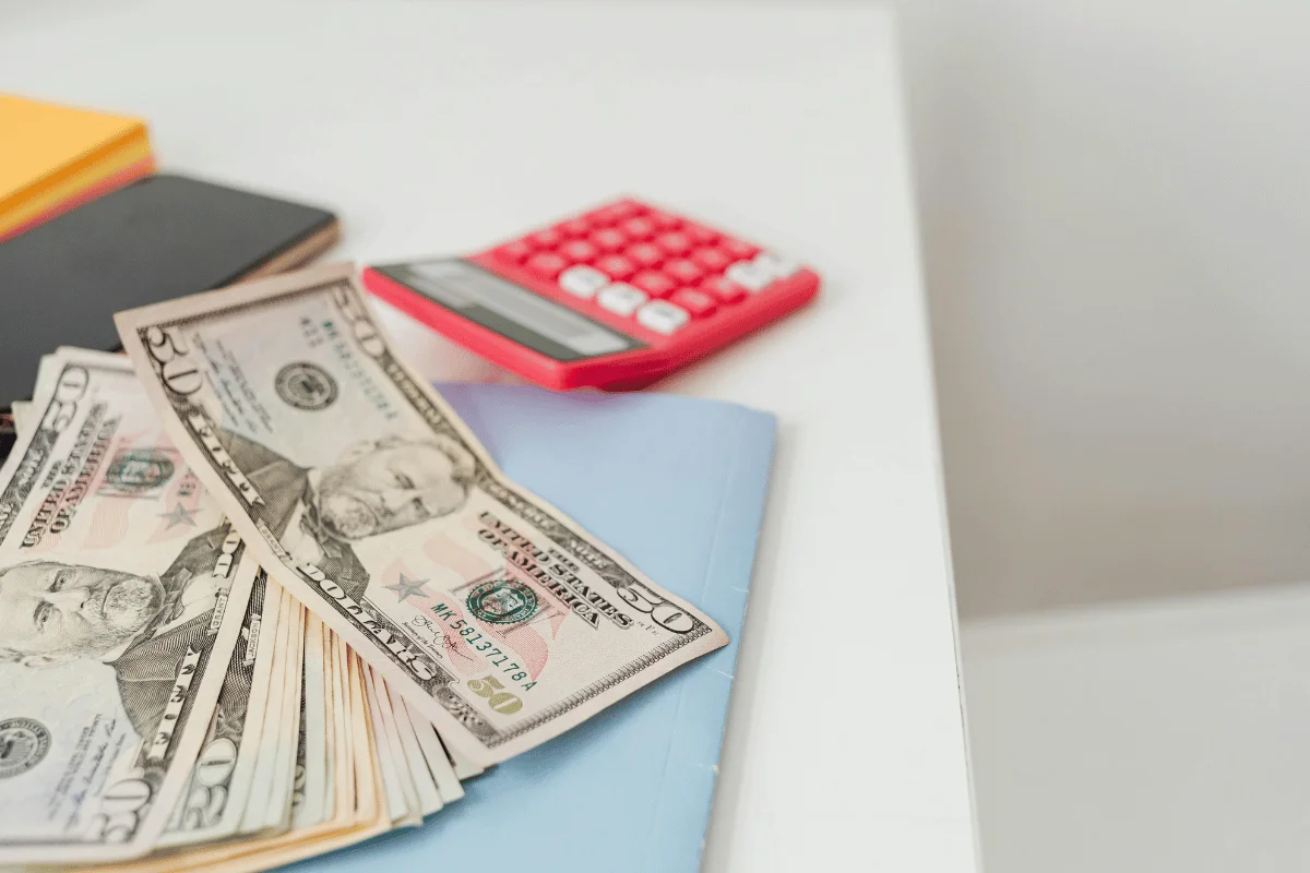 A stack of fifty-dollar bills on a blue folder, with a red calculator in the background on a white table. 
