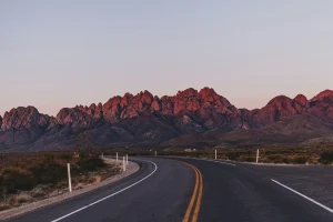 A winding road leads to jagged mountains bathed in a warm, reddish glow at sunset.