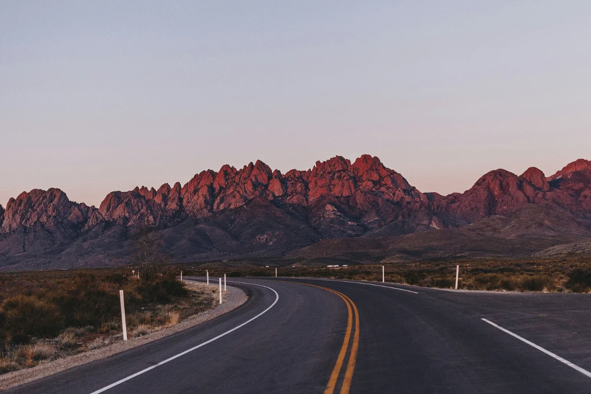 A winding road leads to jagged mountains bathed in a warm, reddish glow at sunset.
