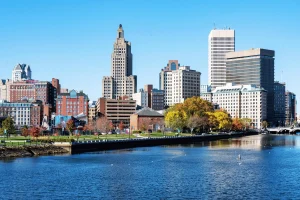 City skyline reflecting in a calm river under a clear blue sky, featuring diverse architectural styles and colorful autumn trees lining the waterfront.