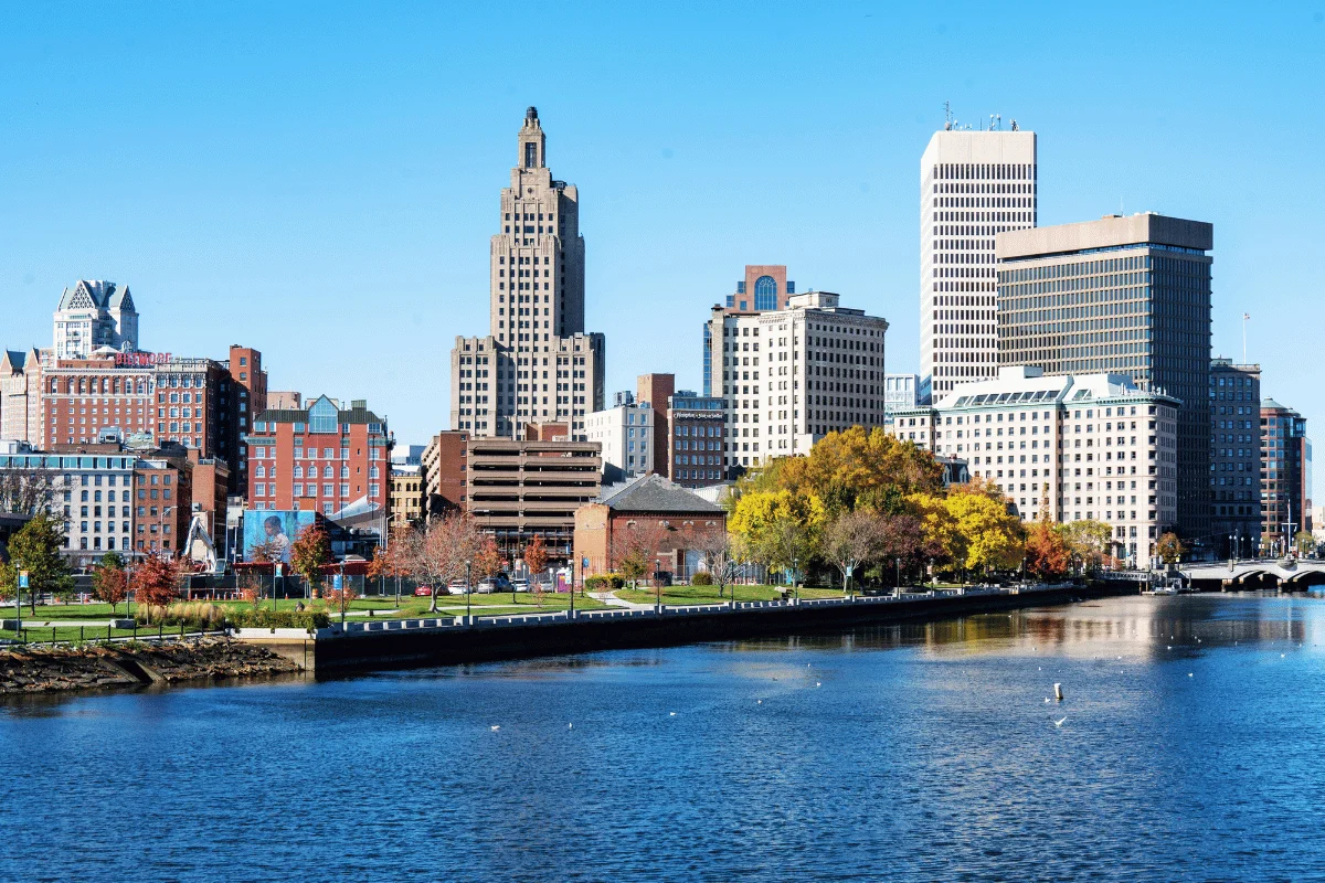 City skyline reflecting in a calm river under a clear blue sky, featuring diverse architectural styles and colorful autumn trees lining the waterfront.