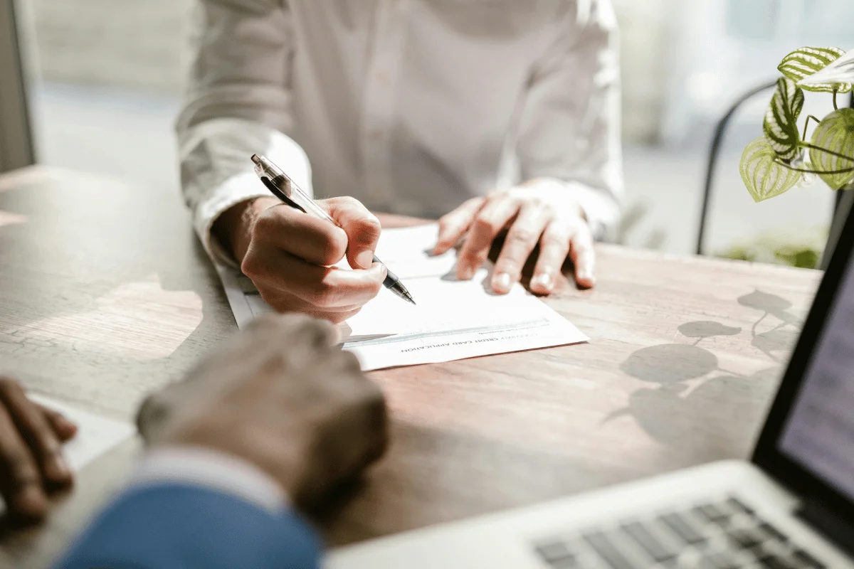 A person in a white shirt signs a document at a sunlit wooden table.