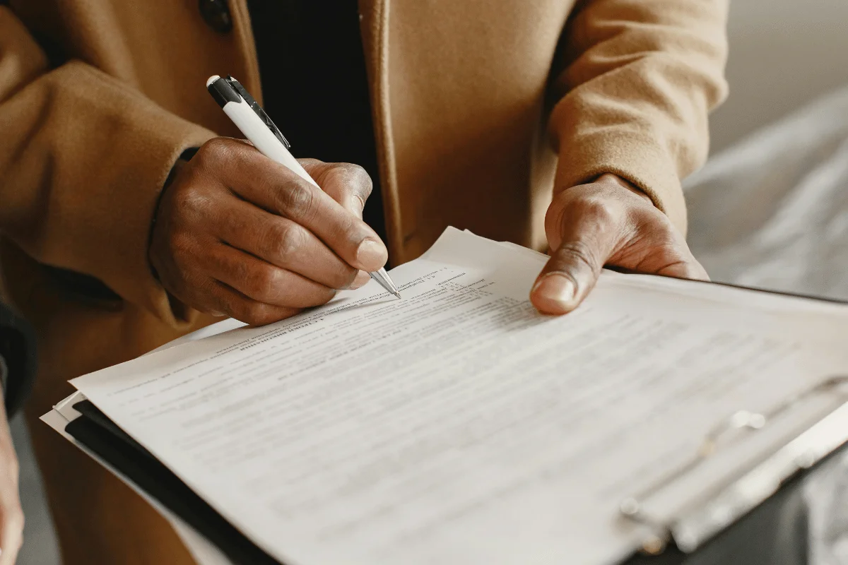 A person wearing a tan coat is holding and signing a document on a clipboard with a pen.