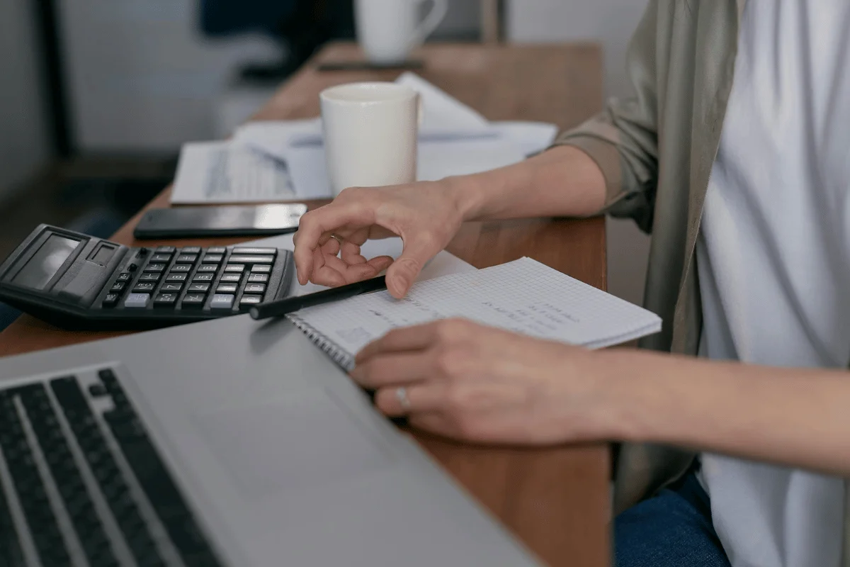 A person works at a wooden desk with a laptop, calculator, and notepad.