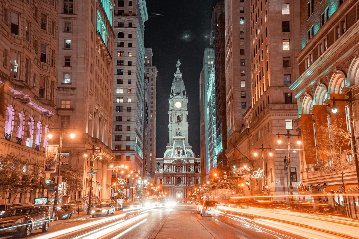 City street at night with tall buildings on either side, leading to an illuminated clock tower. 