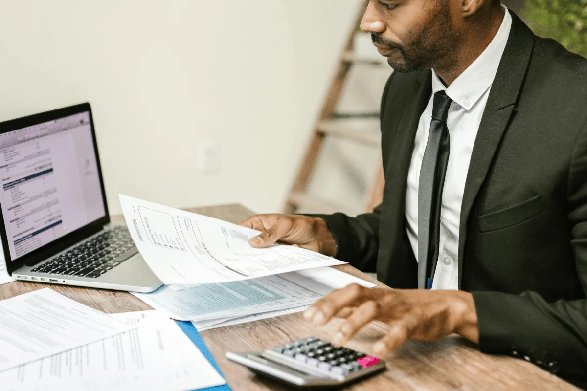 A man in a suit is focused on calculating with a laptop, documents, and calculator on a wooden desk, conveying a professional, busy atmosphere.