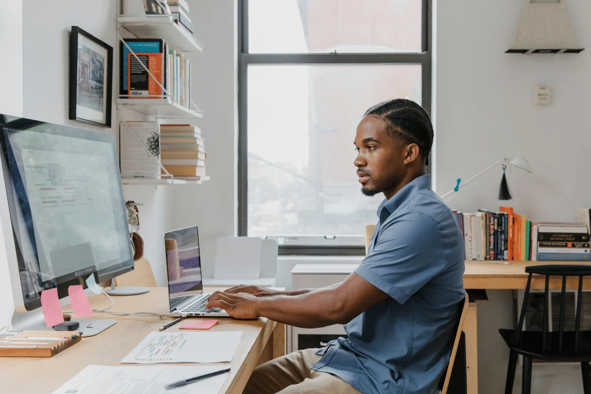 A man in a blue shirt works on a computer at a tidy home office desk, with bookshelves and a large window in the background, conveying focus and productivity.